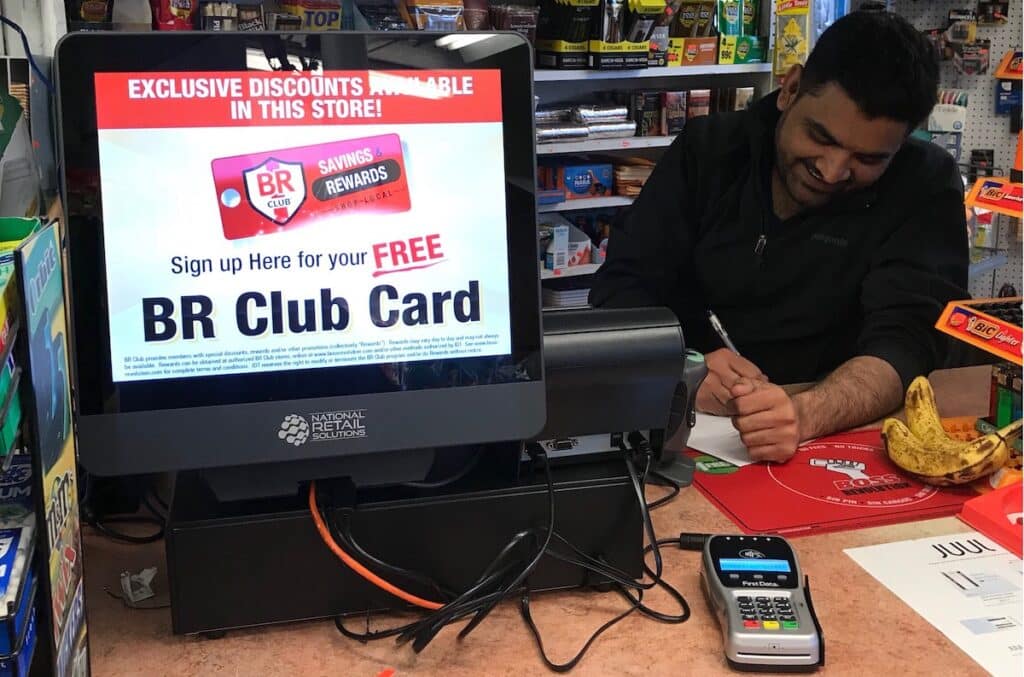 A convenience store owner uses a point of sale hardware with customer facing screen and an EMV card reader. A sales ad for BR Club runs on the screen. Photo by NRS.