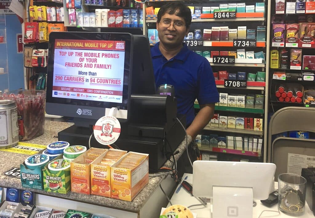 Convenience store owner behind the counter with two different retail point of sale system. Photo by NRS.
