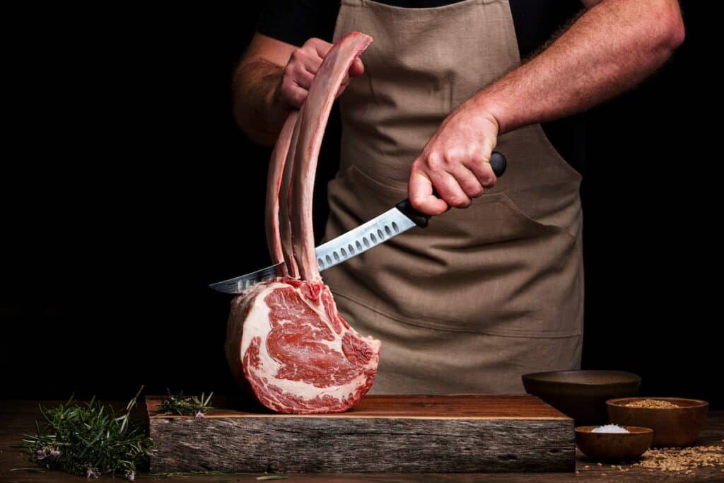 A butcher cuts a large ribeye on the chopping block, with some spices and herbs.