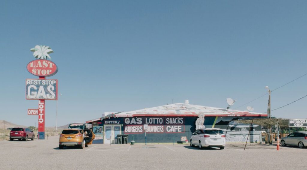 Old gas station in a desert landscape under a clear blue sky, featuring a colorful building with signs for gas, burgers, and souvenirs, several parked cars, and a tall red vintage-style sign with a palm tree on top.