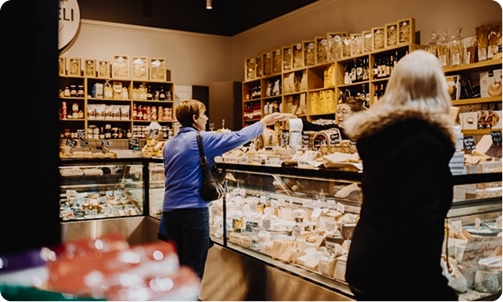Two women shopping at a deli counter with shelves of products in the background