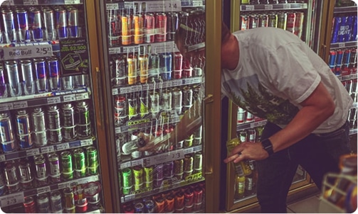 A man in a white t-shirt looks into a glass-fronted refrigerator filled with energy drinks in a store