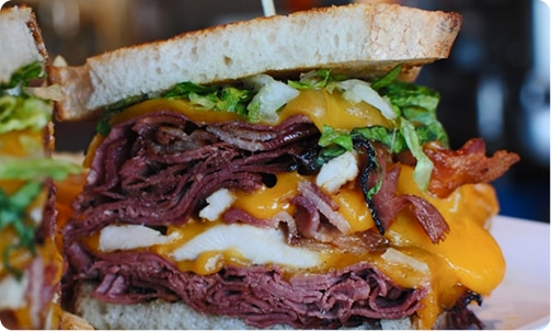 A large, thick sandwich with pastrami, melted cheese, bacon, and lettuce on toasted bread