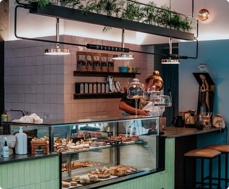 A barista works behind a dessert display case in a cafe with a "DESSERTS" sign overhead
