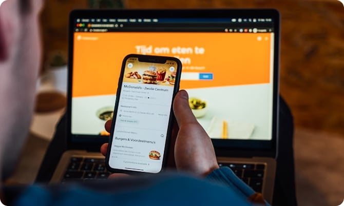 Person holding a smartphone displaying a food delivery app, with a laptop showing a similar website in the background