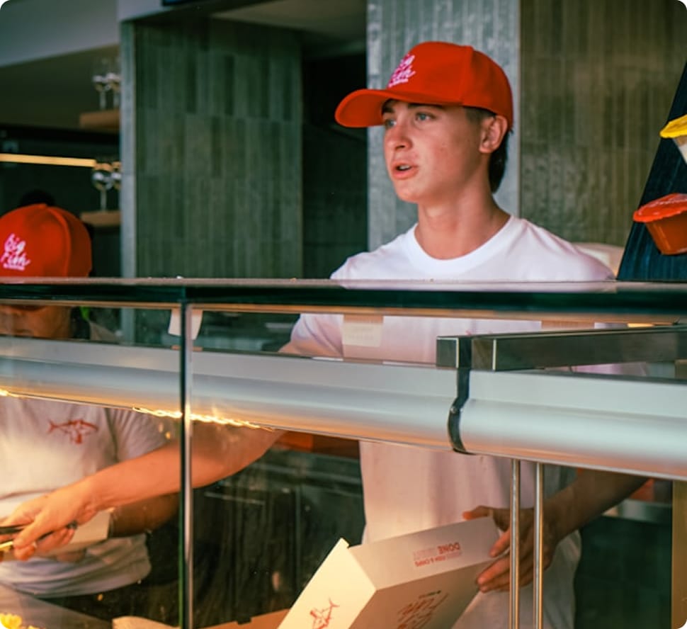 Two people wearing red caps and white shirts, one holding a box, behind a glass counter