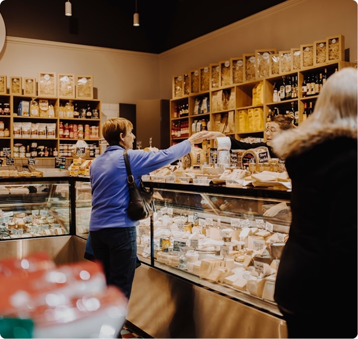 Two women in a cheese shop, one reaching over a display case filled with various cheeses, shelves of products behind them